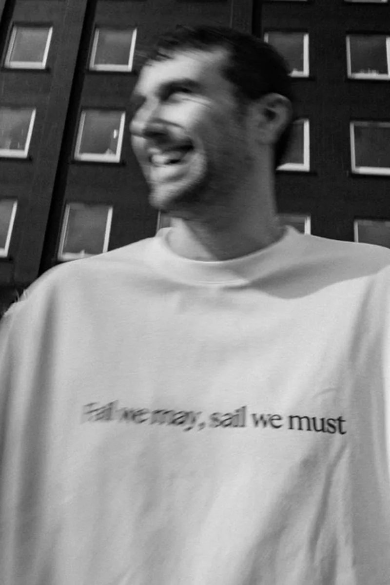 Homme souriant avec un t-shirt 'Eat we may, sail we must' en noir et blanc.