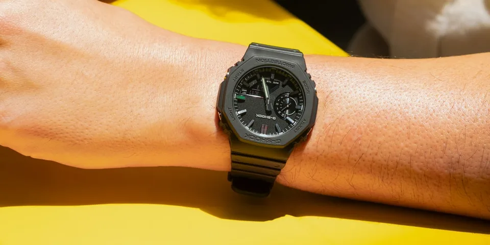 Montre noire au poignet d'une personne sur fond jaune.
