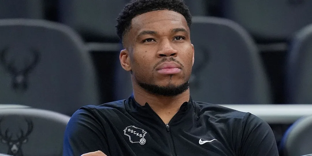 Homme assis avec veste noire NBA, logo Bucks visible.