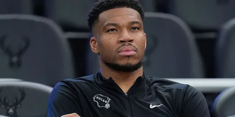 Homme assis avec veste noire NBA, logo Bucks visible.