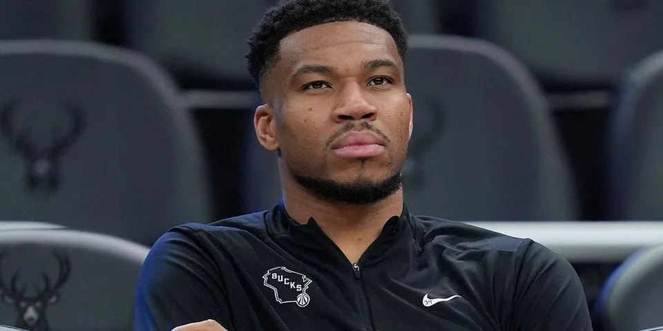 Homme assis avec veste noire NBA, logo Bucks visible.