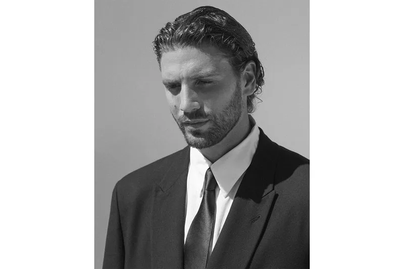 Homme en costume sombre, cheveux lissés en arrière, expression pensive.