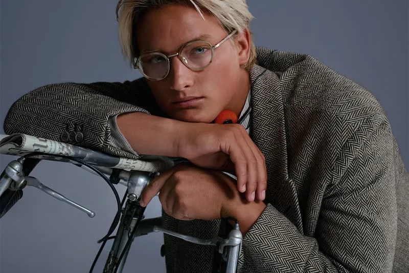 Homme blond avec lunettes appuyé sur un vélo, veste à motif chevron.