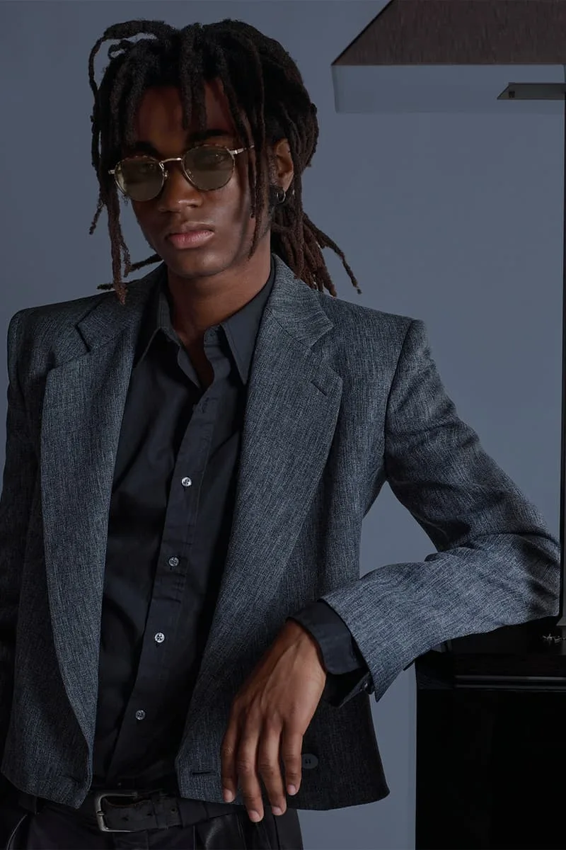 Homme en veste grise avec lunettes de soleil, appuyé sur une lampe.