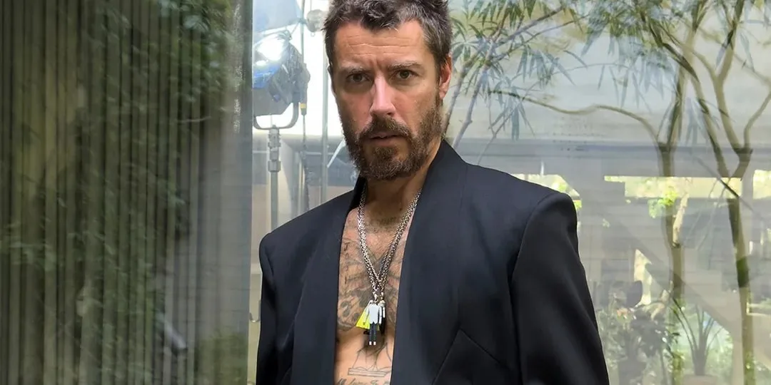 Homme barbu en veste noire, colliers, arrière-plan flou avec plantes