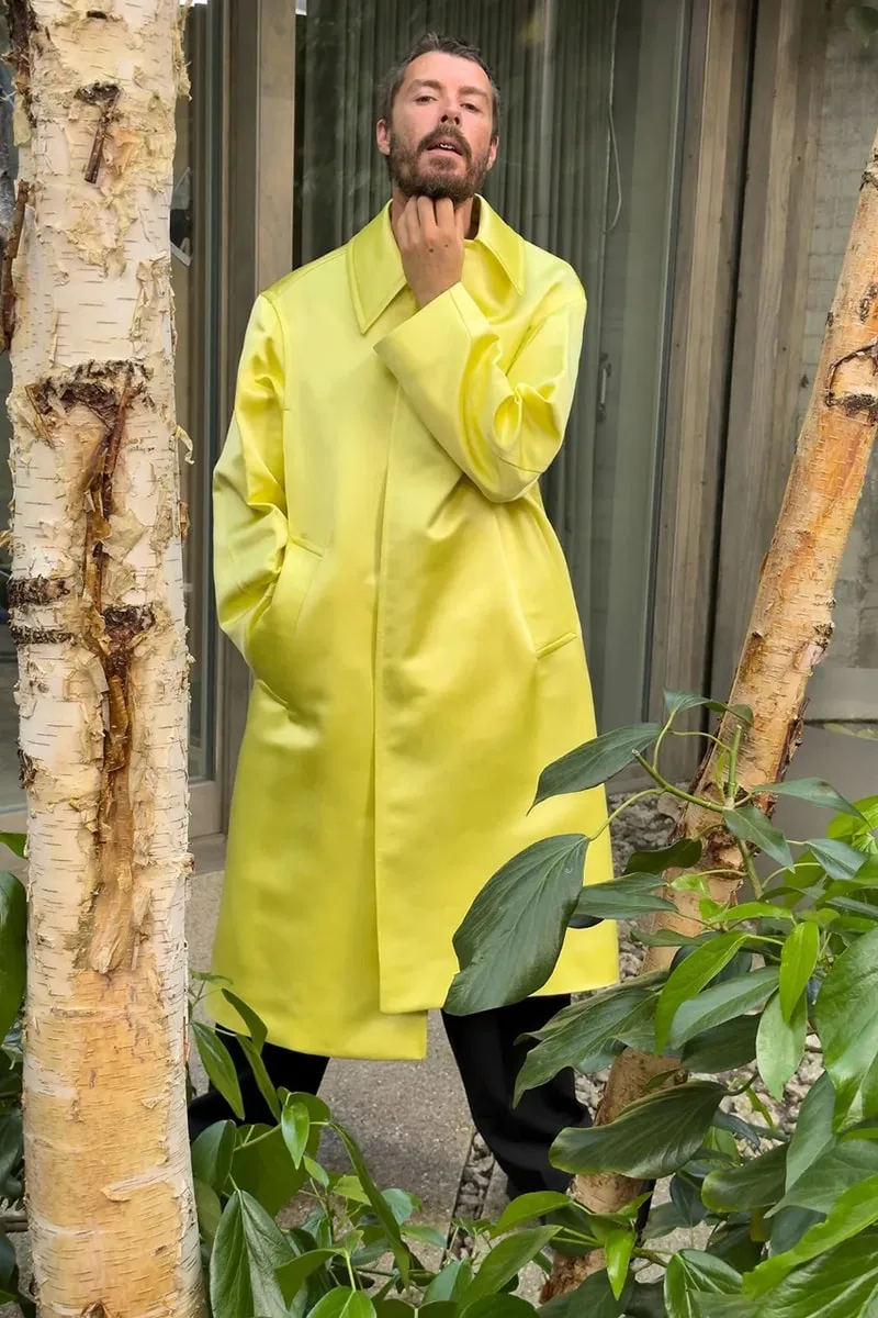 Homme en manteau jaune vif posant dans un jardin.