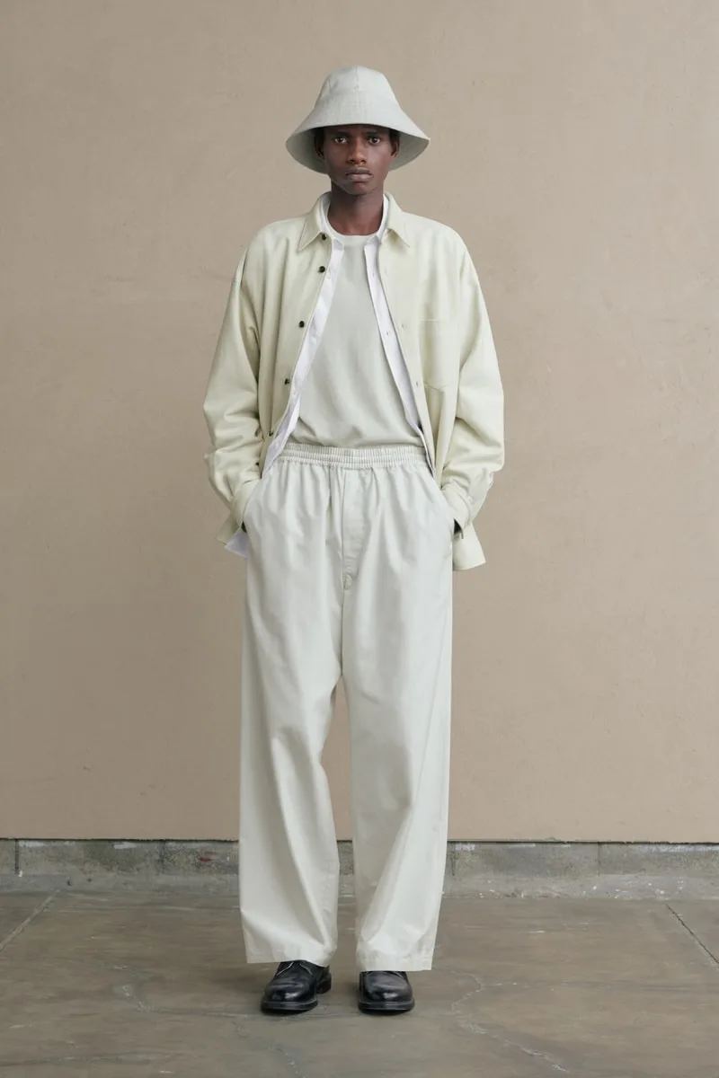 Homme en tenue beige avec chapeau et pantalon ample, posant devant un mur neutre.