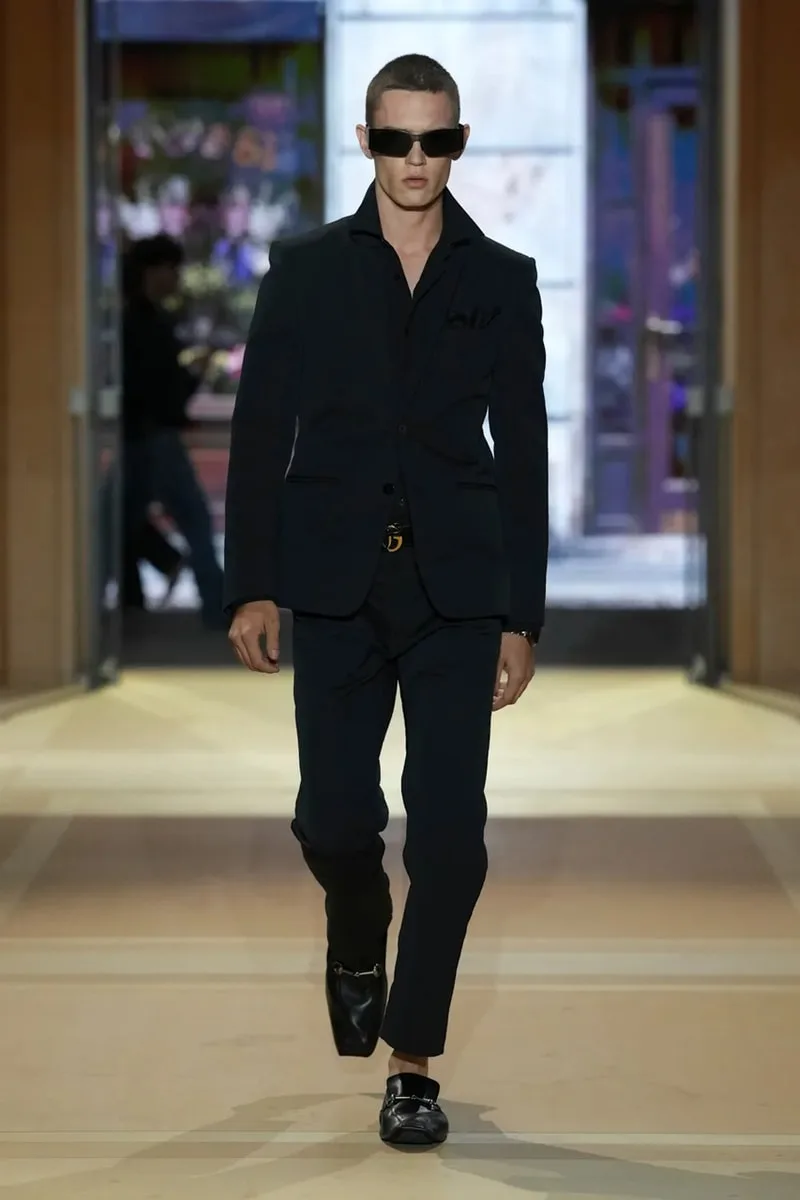 Homme en costume sombre et lunettes de soleil sur un podium, ambiance moderne.
