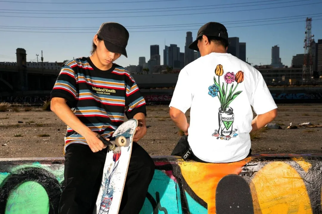 Deux jeunes près d’un mur de graffitis, avec un skateboard et des gratte-ciel en arrière-plan.