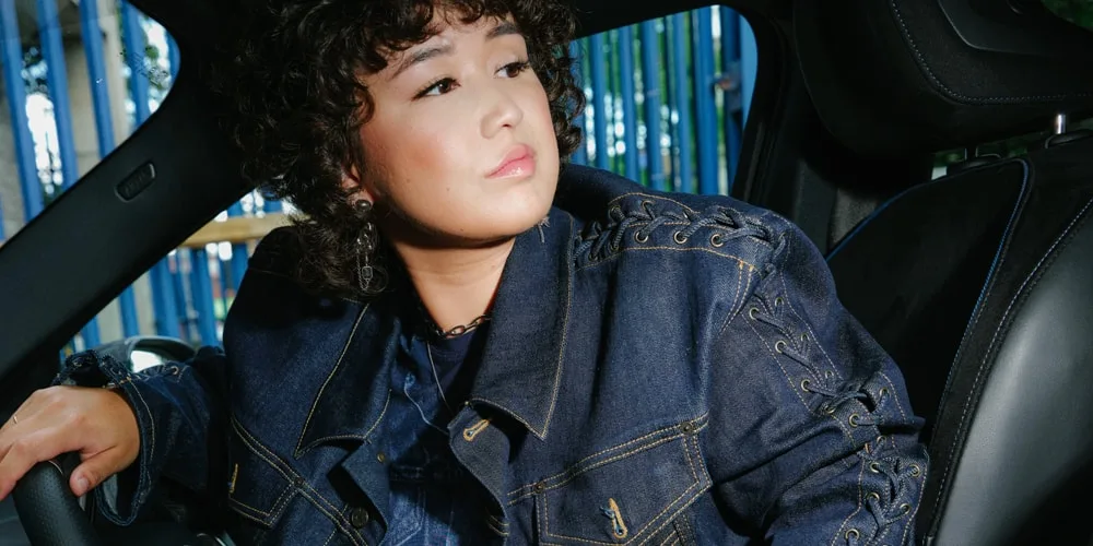 Personne aux cheveux bouclés dans une voiture, portant une veste en jean.