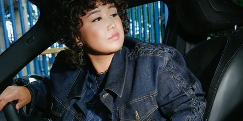 Personne aux cheveux bouclés dans une voiture, portant une veste en jean.