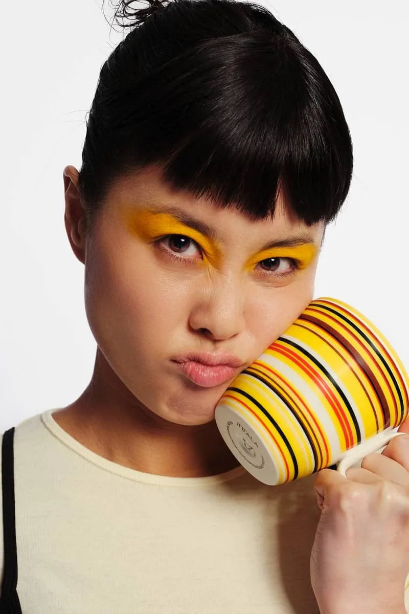 Personne maquillée en jaune tenant une tasse à rayures colorées.