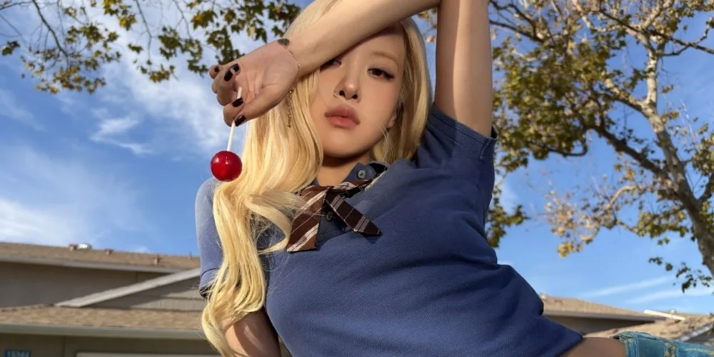 Personne avec cheveux blonds tenant une cerise, ciel bleu et arbre en arrière-plan.