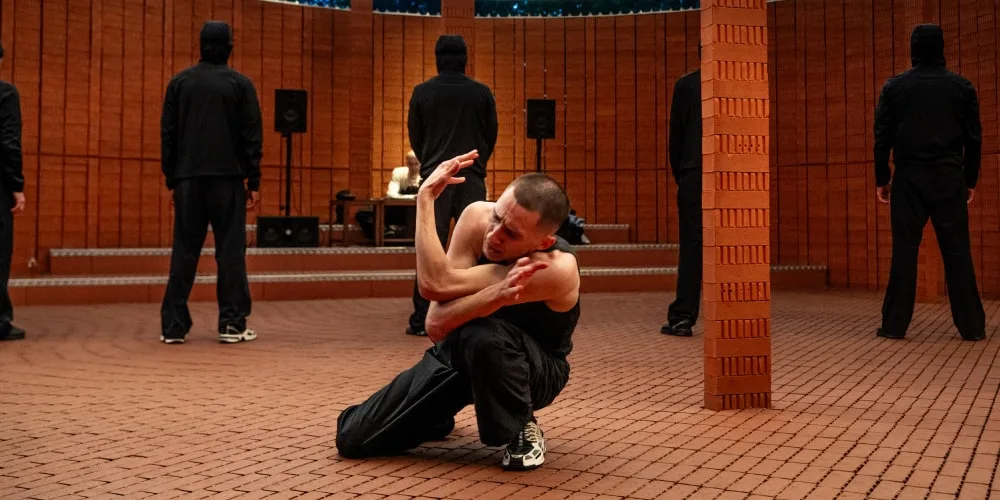 Danseur dans un espace intérieur en briques avec d'autres personnes debout en arrière-plan.