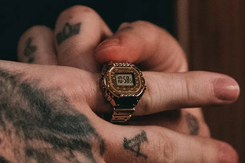 Main tenant une montre Casio dorée sur un doigt avec des tatouages visibles.