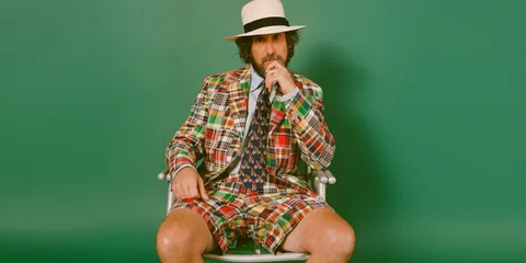 Homme en costume à carreaux colorés, chapeau blanc, fond vert.