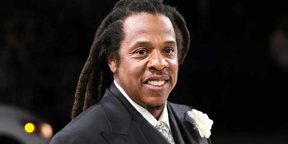 Homme souriant avec des dreadlocks portant un costume sombre et une fleur blanche.
