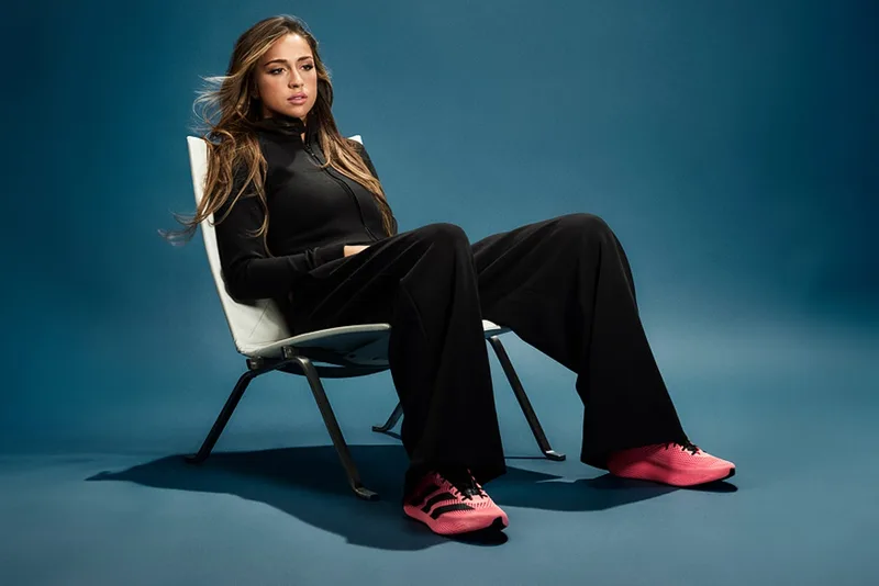 Femme assise avec baskets roses et tenue noire sur fond bleu.