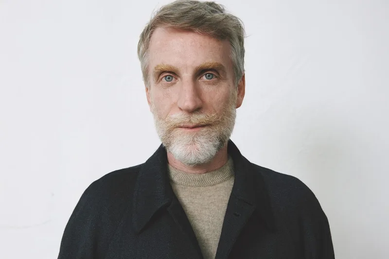 Homme aux cheveux gris et barbe posant sur un fond blanc.