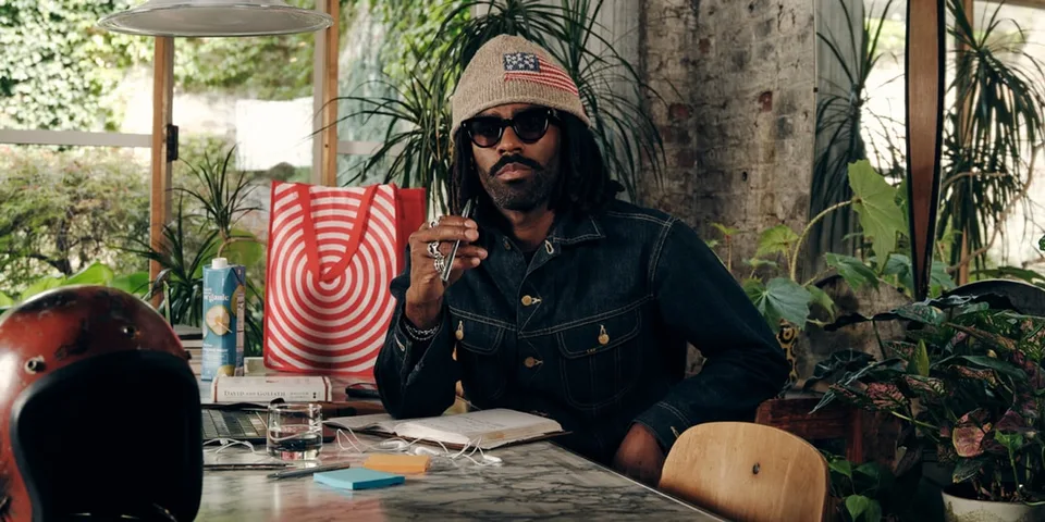 Homme barbu avec lunettes, veste en jean et bonnet assis à une table.