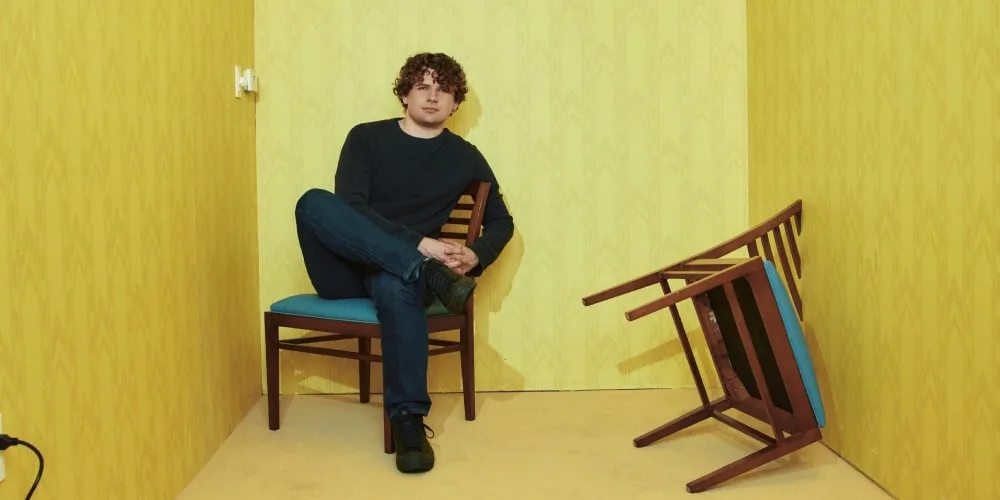 Homme assis sur une chaise dans une pièce jaune, une chaise renversée à côté.