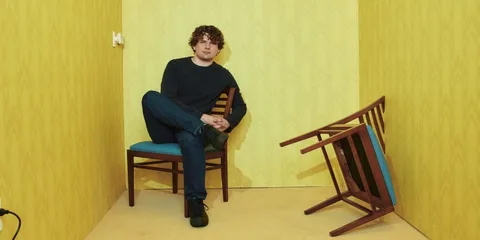 Homme assis sur une chaise dans une pièce jaune, une chaise renversée à côté.