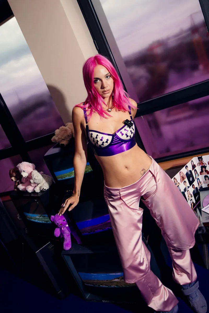 Femme aux cheveux roses portant un haut violet et un pantalon rose.