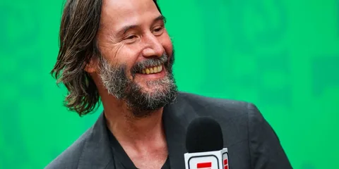 Homme souriant avec micro ESPN, fond vert, cheveux et barbe longs.