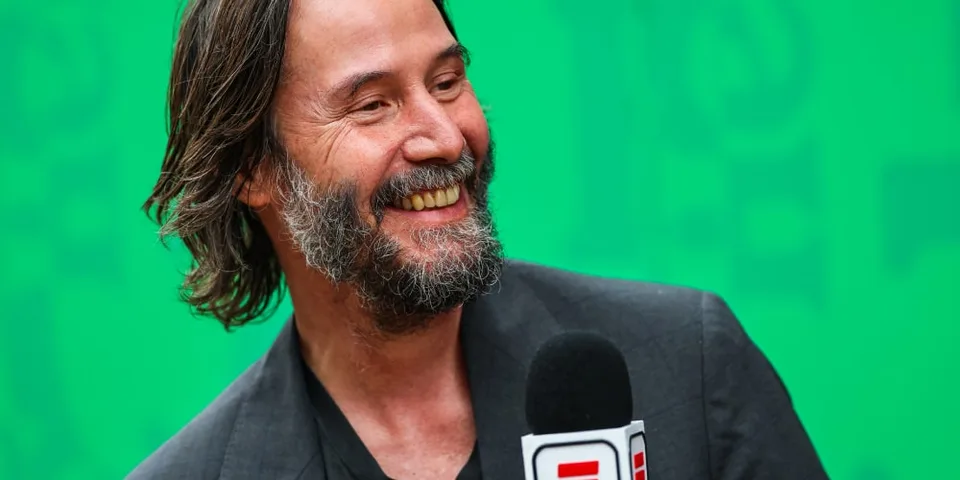 Homme souriant avec micro ESPN, fond vert, cheveux et barbe longs.