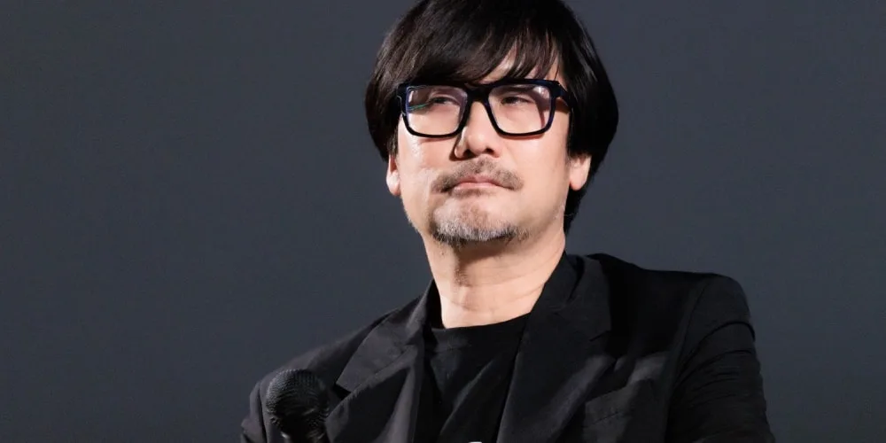 Homme en lunettes noires, cheveux noirs, en costume noir, tient un micro.