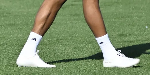 Jambes portant des chaussures Adidas blanches sur gazon.