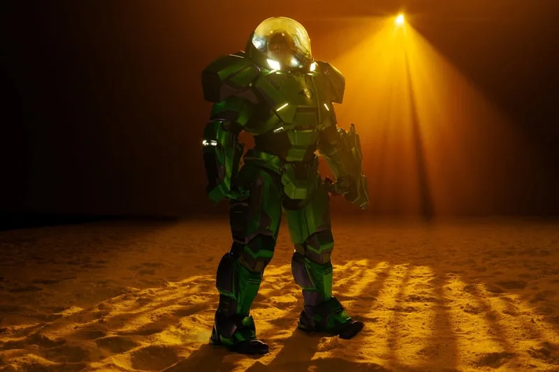 Personne en costume futuriste vert sur sol sablonneux sous éclairage dramatique.