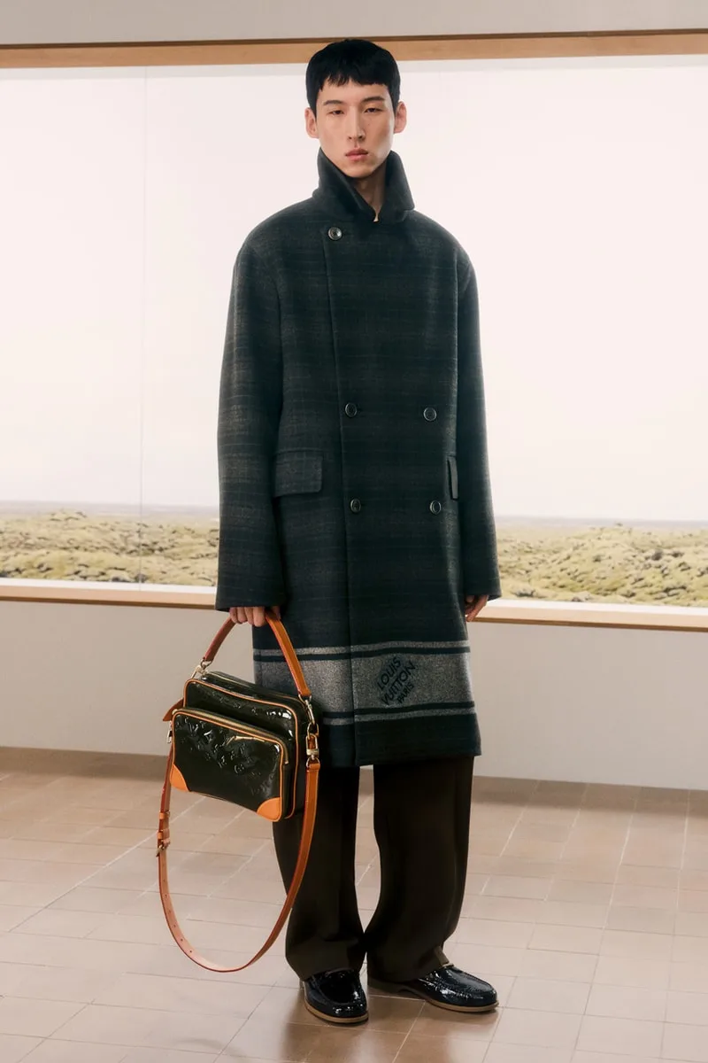 Homme en manteau long et sac à main dans une pièce lumineuse.