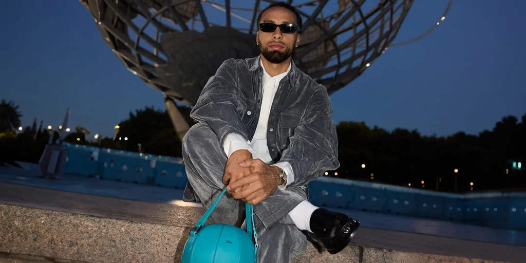 Homme en tenue grise avec sac turquoise devant une grande sculpture métallique.