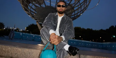 Homme en tenue grise avec sac turquoise devant une grande sculpture métallique.