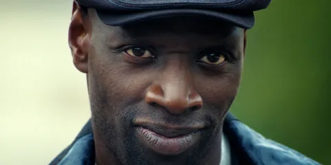 Homme souriant portant une casquette noire, fond flou.