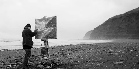 Un peintre réalise un tableau face à une plage sauvage et aux falaises.