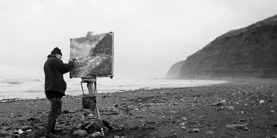 Un peintre réalise un tableau face à une plage sauvage et aux falaises.