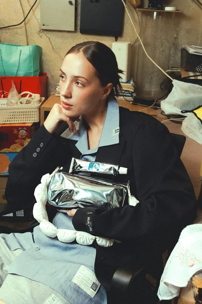 Femme assise, veste noire, sac argenté et tube, environnement de bureau désordonné.