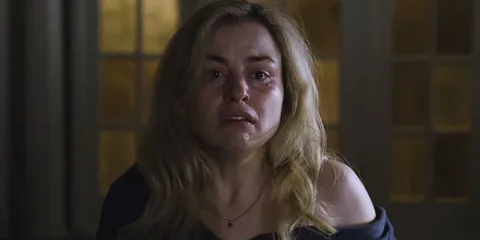 Femme blonde en larmes, assise dans une pièce sombre.