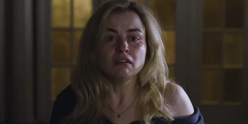 Femme blonde en larmes, assise dans une pièce sombre.