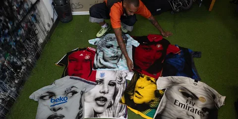 Homme avec t-shirts peints, visage de célébrités, sur un sol vert.