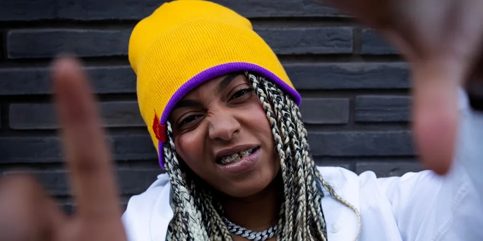 Personne portant un bonnet jaune et des tresses, posant devant un mur sombre.