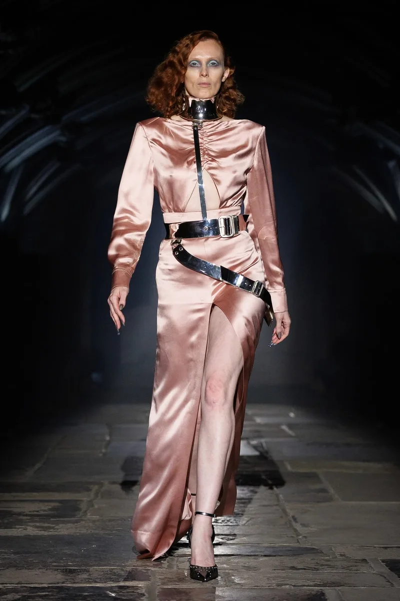 Robe en satin rose avec accessoires métalliques sur un podium sombre.