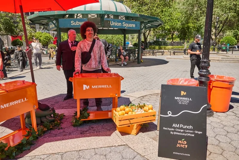Personne avec stand de boissons, chariots oranges, place publique en arrière-plan.