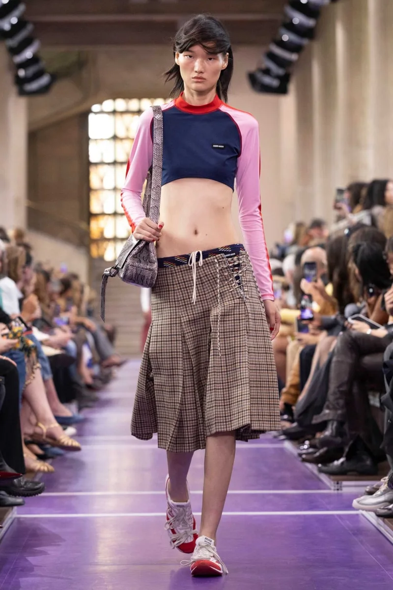 Défilé de mode avec mannequin portant un haut coloré et une jupe à carreaux.