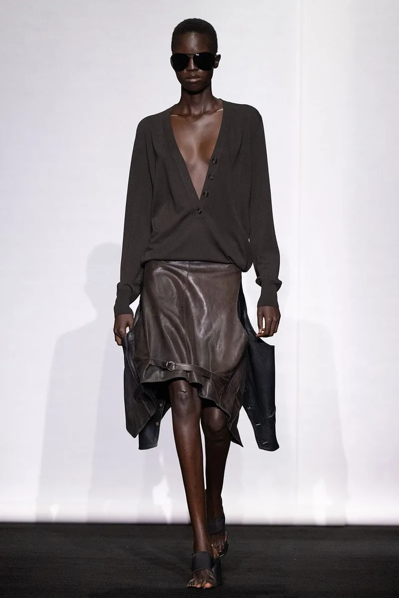 Défilé de mode avec mannequin en tenue sombre et lunettes, jupe en cuir fluide.