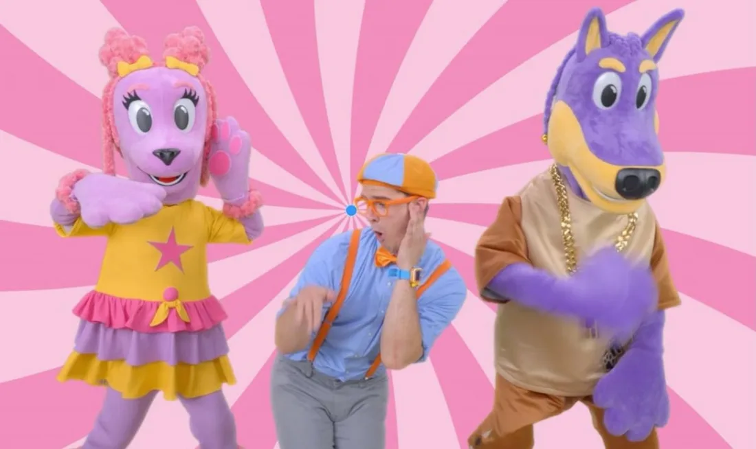 Personnage en costume coloré avec deux mascottes sur fond rose.