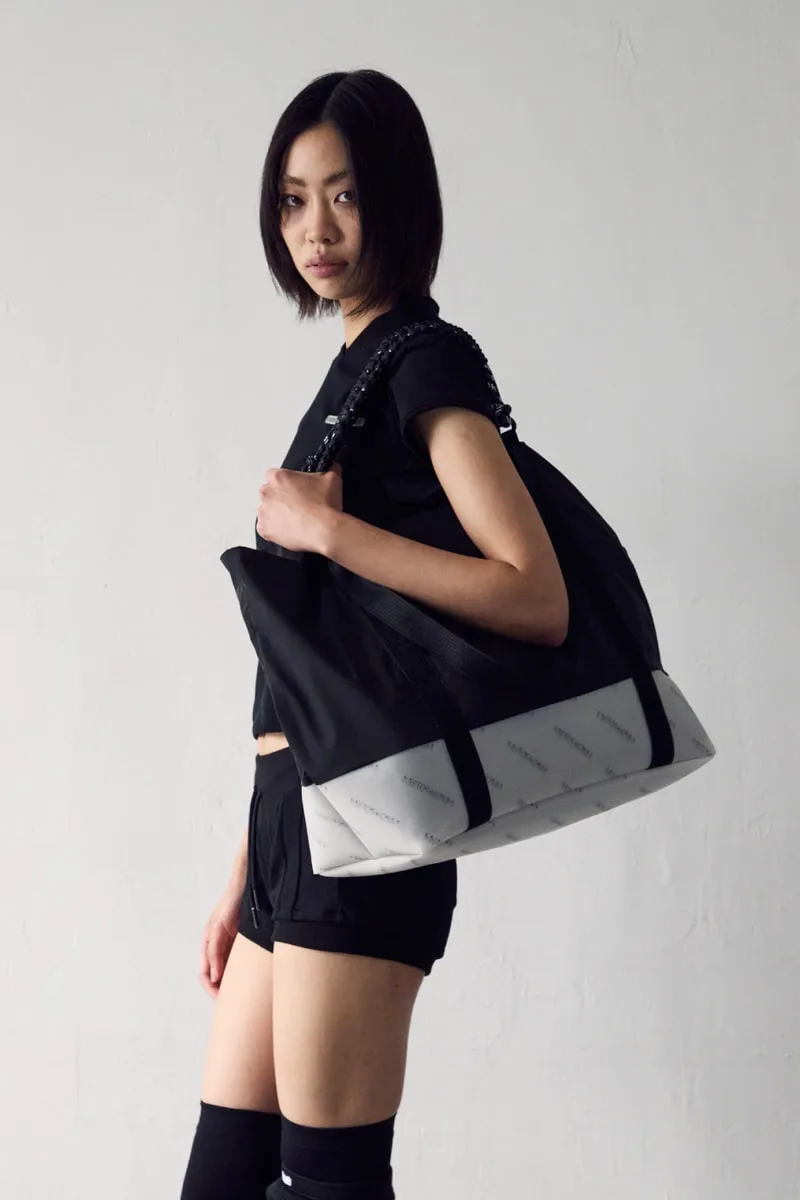 Personne avec un grand sac noir et blanc, en tenue noire, posant sur fond neutre.