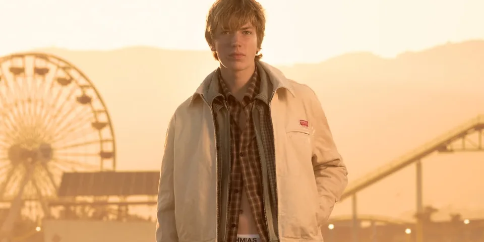 Jeune homme en veste beige devant une grande roue au coucher du soleil.